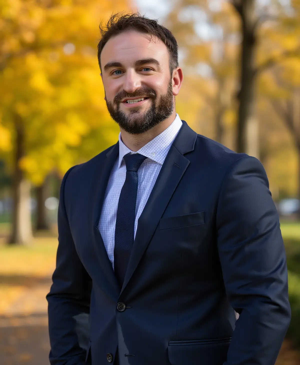 A smiling professional in a navy suit standing outdoors among autumn trees